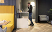 Person using a laptop standing at adjustable desk in a modern office setting with yellow booth and gray chair.