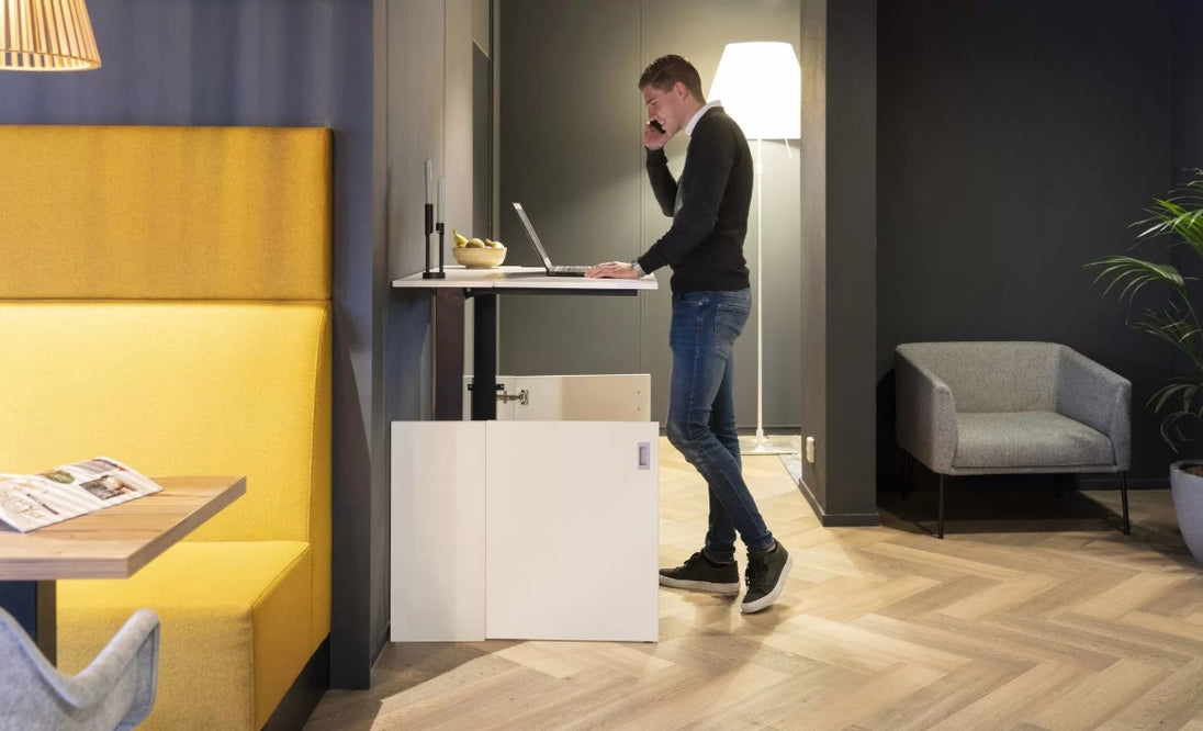 Person using a laptop standing at adjustable desk in a modern office setting with yellow booth and gray chair.