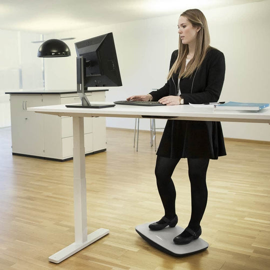 A person standing on a gray balance board that is placed under a desk, using a computer setup on the desk.