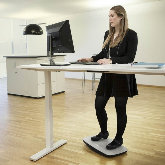 A person standing on a gray balance board that is placed under a desk, using a computer setup on the desk.